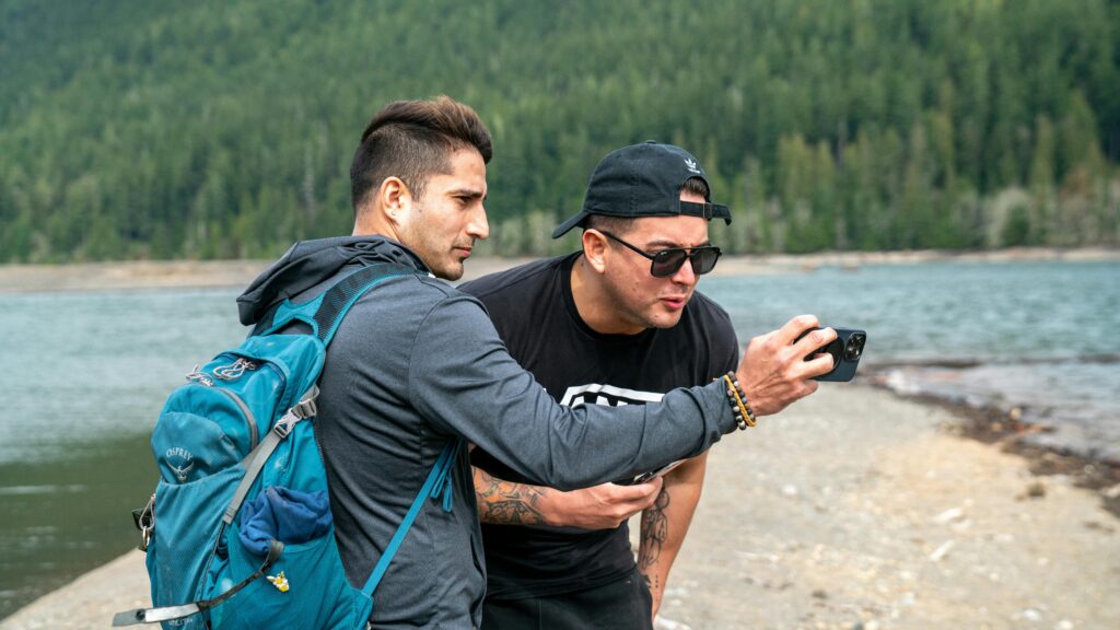 Two men enjoy a scenic outdoor adventure by the lake, capturing the moment with a smartphone.